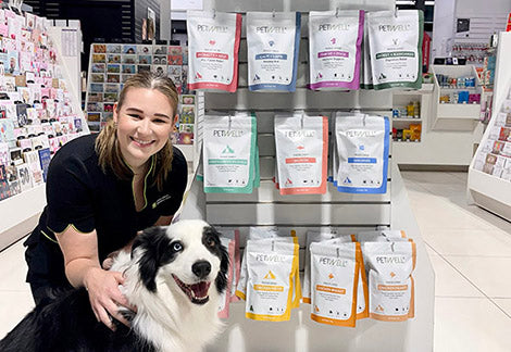 Pharmacy assistant crouching with border collie next to PetWell treat display in store